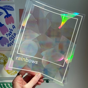 May include: Clear, iridescent sticker sheets with a geometric design and the word "rainbows" in a holographic font. The stickers have a square border and are held up against a white wall.