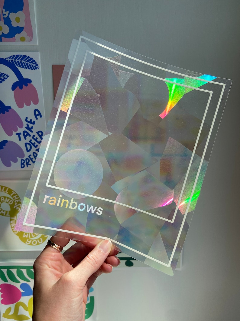May include: Clear, iridescent sticker sheets with a geometric design and the word "rainbows" in a holographic font. The stickers have a square border and are held up against a white wall.