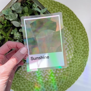 May include: A rectangular sticker with a holographic finish that reflects rainbow colors. The sticker features a white border and a white rectangular section at the bottom containing the black printed text Sunshine in a sans-serif font.