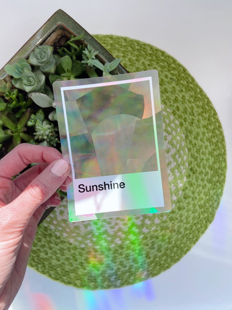 May include: A rectangular sticker with a holographic finish that reflects rainbow colors. The sticker features a white border and a white rectangular section at the bottom containing the black printed text Sunshine in a sans-serif font.
