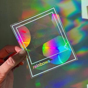 May include: A clear, rectangular sticker with a white border and the word "rainbows" in the bottom left corner. The sticker features a holographic design with rainbow colors and geometric shapes.