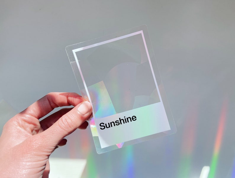 May include: A rectangular, transparent sticker featuring a white border and a white rectangular block at the bottom with the word Sunshine printed in black. The sticker has a holographic finish that reflects rainbow colors when light hits it.