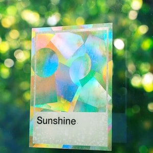 May include: A rectangular holographic sticker featuring a geometric pattern of overlapping circles and squares in iridescent colors. The sticker has a white border and a white rectangular section at the bottom with the word Sunshine printed in black.