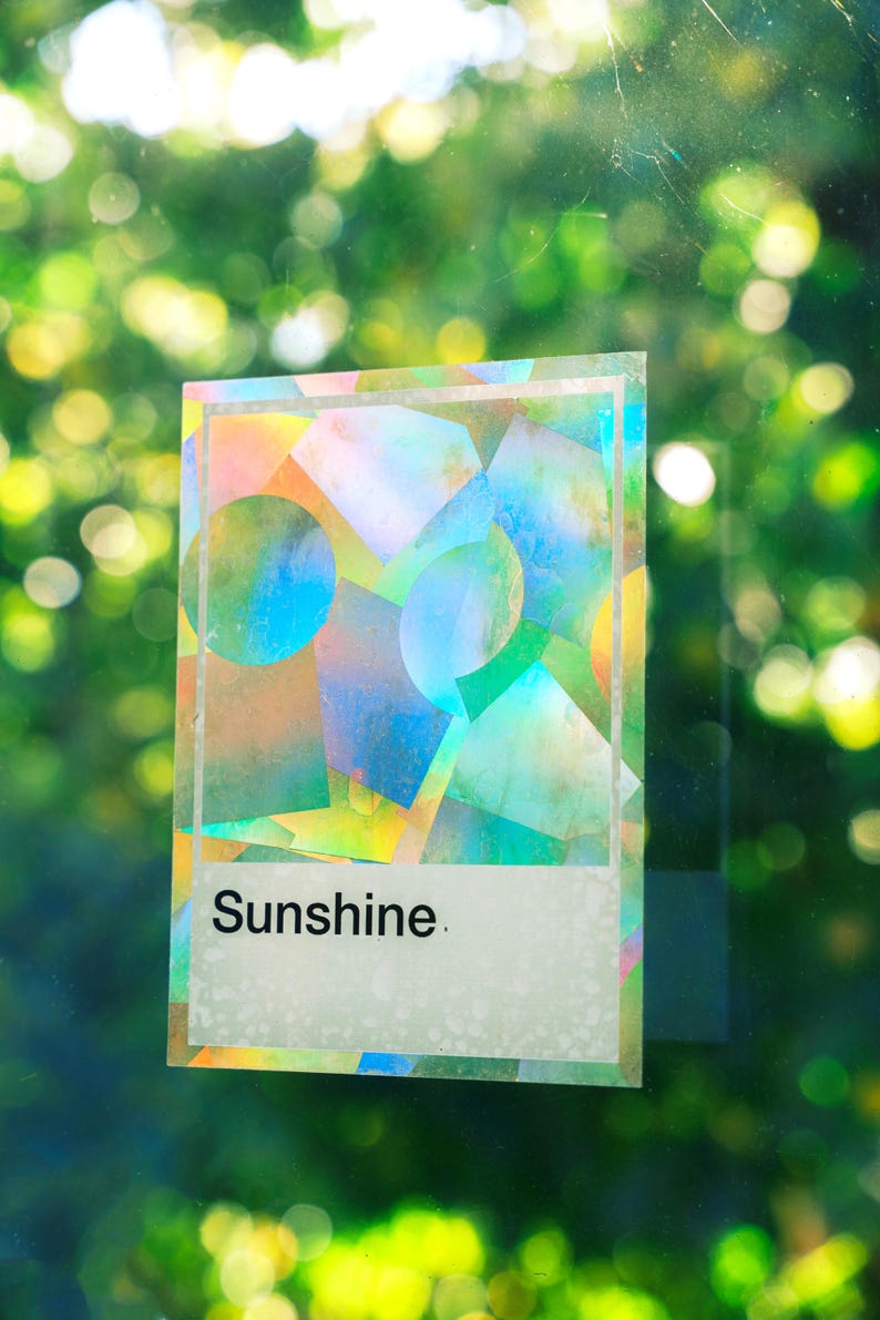May include: A rectangular holographic sticker featuring a geometric pattern of overlapping circles and squares in iridescent colors. The sticker has a white border and a white rectangular section at the bottom with the word Sunshine printed in black.