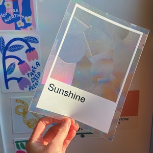 May include: A rectangular, holographic, semi-transparent sticker featuring a white border and a white rectangular base at the bottom. The word "Sunshine" is printed in black, sans-serif text on the white base. The sticker has a glittery finish.