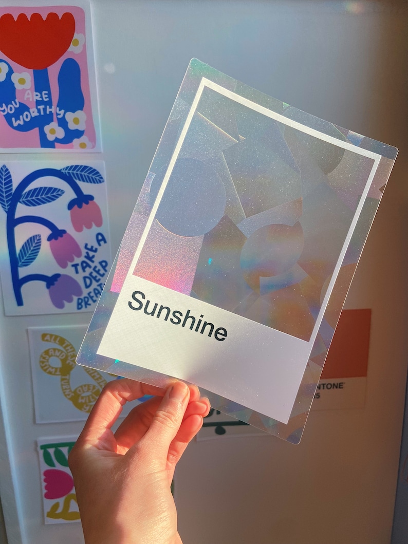 May include: A rectangular, holographic, semi-transparent sticker featuring a white border and a white rectangular base at the bottom. The word "Sunshine" is printed in black, sans-serif text on the white base. The sticker has a glittery finish.