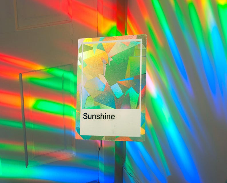 May include: A rectangular card featuring a holographic, multi-faceted geometric pattern in shades of green, yellow, and blue. The lower portion contains a white rectangular block with the word Sunshine printed in bold, black, sans-serif text.