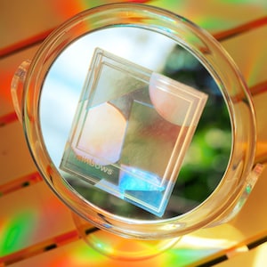 May include: A clear, circular magnifying glass with a handle, holding a transparent, rectangular object with the word "rainbows" printed on it. The object has geometric shapes and reflects rainbow colors.