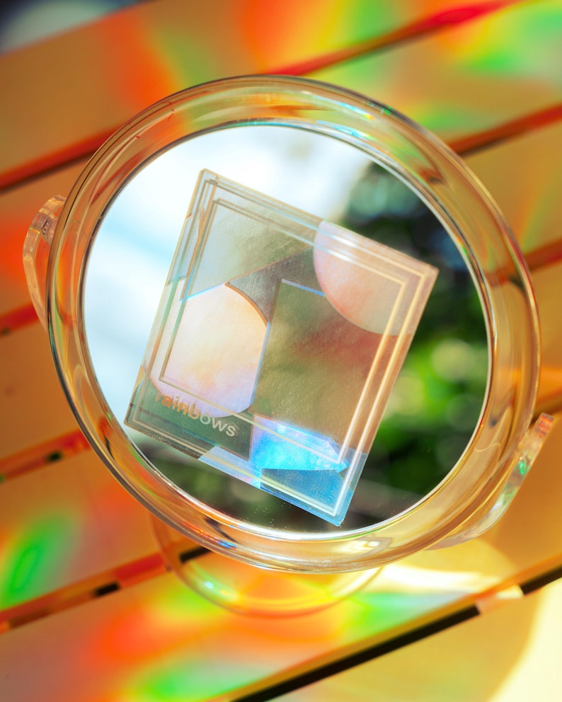 May include: A clear, circular magnifying glass with a handle, holding a transparent, rectangular object with the word "rainbows" printed on it. The object has geometric shapes and reflects rainbow colors.