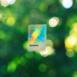 May include: A rectangular sticker with a holographic finish featuring geometric shapes in various colors. The sticker has a white rectangular section at the bottom with the word Sunshine printed in black text, centered on a clear glass surface.