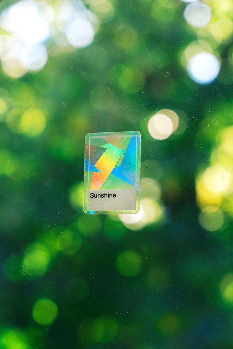 May include: A rectangular sticker with a holographic finish featuring geometric shapes in various colors. The sticker has a white rectangular section at the bottom with the word Sunshine printed in black text, centered on a clear glass surface.