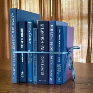 May include: A stack of seven hardcover books in various shades of blue, tied together with a light blue ribbon. The spines display titles including Manchester, Impact Players, Jimmy Carter, Atlantis Found, Field of Hope, and Chili Dawgs Always Bark at Night.