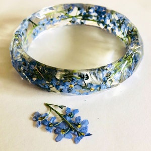 May include: A clear resin bangle bracelet with blue forget-me-not flowers embedded inside. A small cluster of forget-me-not flowers is also shown separately.
