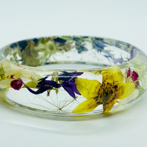May include: A clear resin bangle bracelet with embedded dried flowers. The bracelet features a vibrant mix of yellow, purple, white, and red flowers, creating a unique and colorful accessory. The bangle is a circular shape.