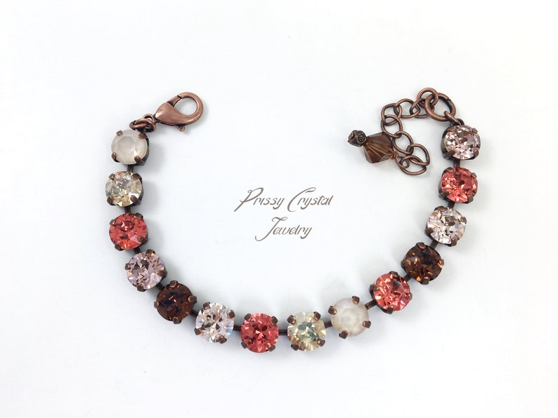 May include: A copper-toned bracelet featuring a series of colorful crystals in shades of pink, peach, brown, and cream. The bracelet has a lobster clasp closure and an adjustable chain. The text "Prissy Crystal Jewelry" is visible.