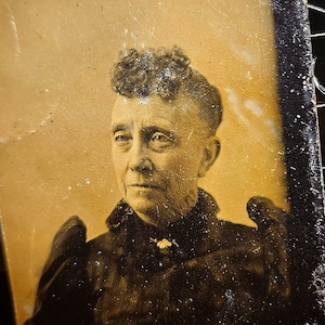 Może przedstawiać: Antyczny portret kobiety w czarnej sukni, prezentowany w prostokątnej ramie. Obraz w odcieniach sepii przedstawia kobietę z poważnym wyrazem twarzy. Rama jest lekko uszkodzona, co dodaje uroku vintage. Tło jest ciemne.