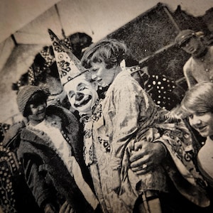 May include: A black and white photo of a clown with a large, colorful hat, interacting with a group of children. The clown is smiling and holding a child's hand. The children are looking at the clown with smiles on their faces.