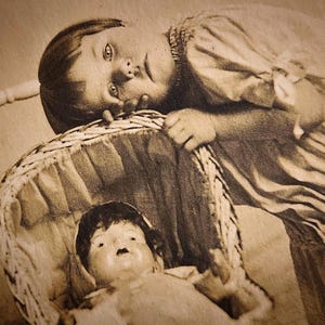 Peut inclure: Photographie sépia d'une enfant penchée sur un berceau en osier contenant une poupée. L'enfant porte une robe claire et semble regarder la poupée. La poupée a les cheveux foncés et porte une tenue claire.