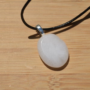Peut inclure: Un collier avec un pendentif blanc ovale, une b&eacute;li&egrave;re argent&eacute;e et un cordon noir. Le pendentif est lisse et opaque, et le collier est pos&eacute; sur une surface en bois brun clair. Le pendentif mesure environ 4 cm de long.
