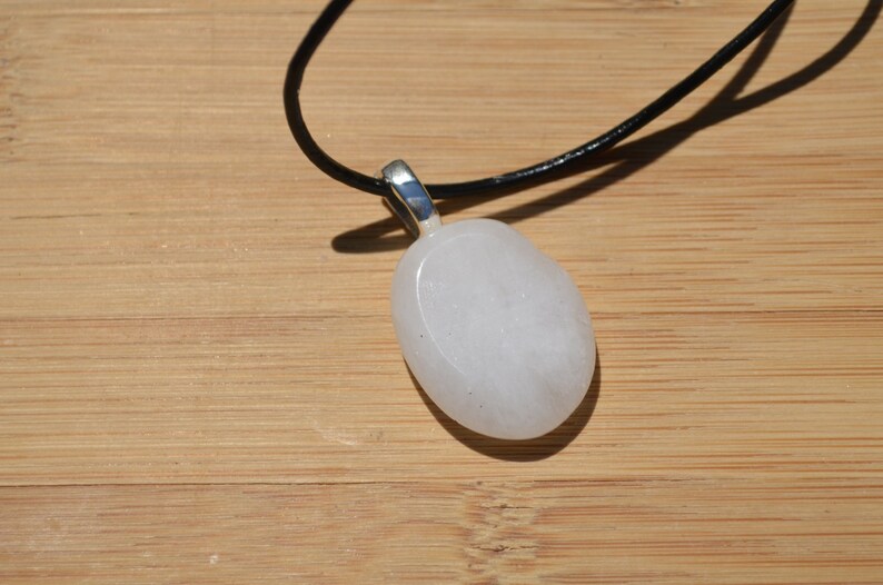 Peut inclure: Un collier avec un pendentif blanc ovale, une b&eacute;li&egrave;re argent&eacute;e et un cordon noir. Le pendentif est lisse et opaque, et le collier est pos&eacute; sur une surface en bois brun clair. Le pendentif mesure environ 4 cm de long.