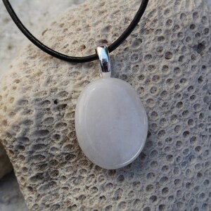Peut inclure: Un collier avec un pendentif ovale blanc et une b&eacute;li&egrave;re argent&eacute;e. Le pendentif est suspendu &agrave; un cordon noir. Le collier repose sur une surface textur&eacute;e et claire, peut-&ecirc;tre du corail ou de la pierre.