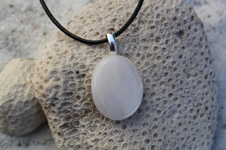 Peut inclure: Un collier avec un pendentif ovale blanc et une b&eacute;li&egrave;re argent&eacute;e. Le pendentif est suspendu &agrave; un cordon noir. Le collier repose sur une surface textur&eacute;e et claire, peut-&ecirc;tre du corail ou de la pierre.
