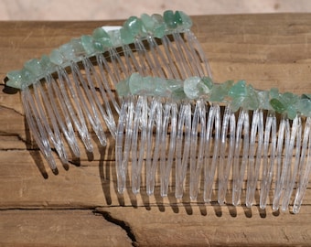 May include: Two clear plastic hair combs, each adorned with a row of small, light green gemstones. The combs are positioned on a weathered, brown wooden surface, with the sunlight casting shadows.