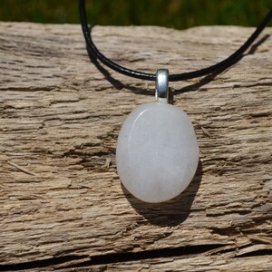 Peut inclure: Un collier avec un pendentif blanc de forme ovale, une b&eacute;li&egrave;re argent&eacute;e et un cordon noir. Le pendentif est lisse et opaque. Le collier est pr&eacute;sent&eacute; sur une surface en bois brun vieilli.
