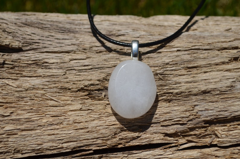 Peut inclure: Un collier avec un pendentif blanc de forme ovale, une b&eacute;li&egrave;re argent&eacute;e et un cordon noir. Le pendentif est lisse et opaque. Le collier est pr&eacute;sent&eacute; sur une surface en bois brun vieilli.