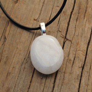 Peut inclure: Un collier avec un pendentif blanc ovale, une b&eacute;li&egrave;re argent&eacute;e et un cordon noir. Le pendentif est lisse et brillant. Le collier est pos&eacute; sur une surface en bois vieilli.