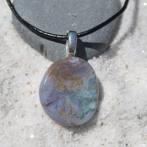 May include: A pendant necklace featuring a polished, oval-shaped stone with a mix of blue, green, and brown hues. The stone is suspended from a silver-colored bail on a black cord, set against a backdrop of white sand.