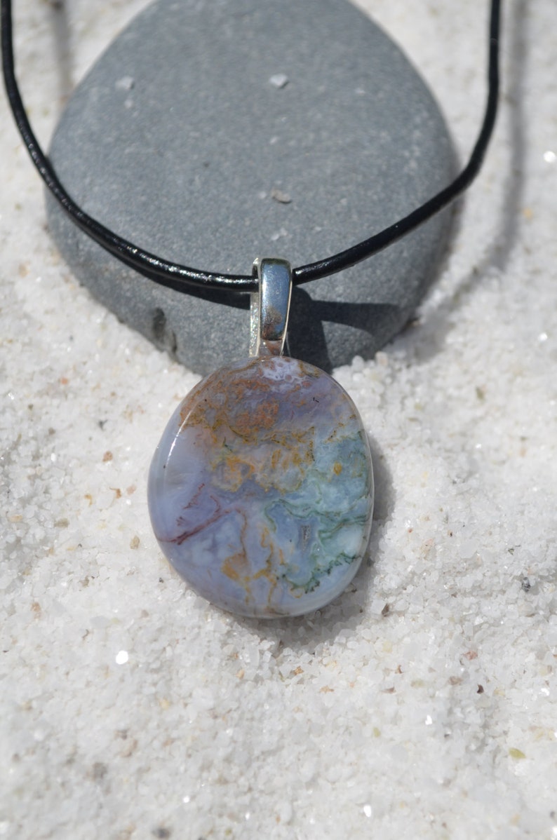 May include: A pendant necklace featuring a polished, oval-shaped stone with a mix of blue, green, and brown hues. The stone is suspended from a silver-colored bail on a black cord, set against a backdrop of white sand.
