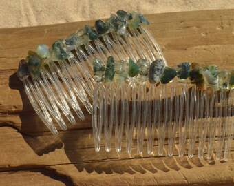 May include: Two clear plastic hair combs are adorned with green and white moss agate stones. The stones are irregularly shaped and set along the top edge of the combs. The combs are displayed on a wooden surface.