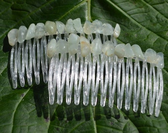 May include: Two clear plastic hair combs, each adorned with a row of pale green, irregular-shaped stones. The combs are set against a dark green leaf, creating a natural and elegant aesthetic. These hair accessories are ideal for adding a touch of nature-inspired beauty.