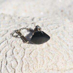 Op de afbeelding: Een zwarte, druppelvormige flesstopper met een zilveren ketting en een T-bar. De stopper is gemaakt van een glanzend, donker materiaal en staat tegen een getextureerde, witte achtergrond.