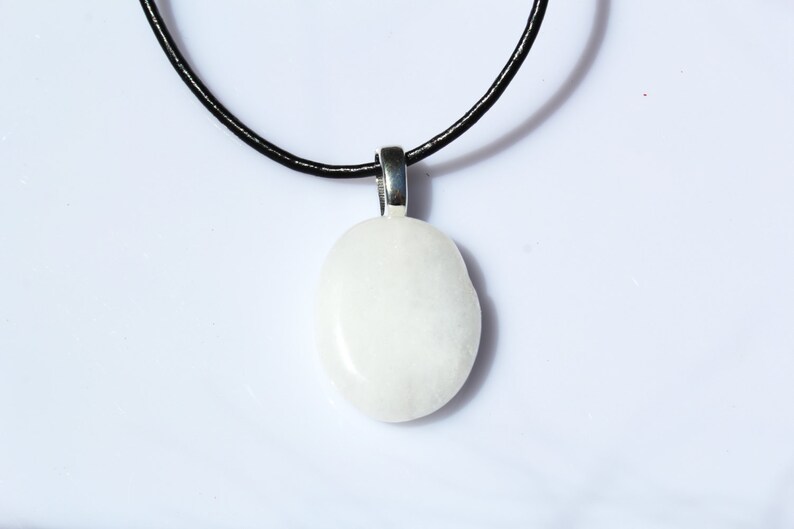 Peut inclure: Un collier avec un pendentif blanc ovale. Le pendentif est suspendu &agrave; un cordon noir. Le pendentif est fait d'un mat&eacute;riau lisse et opaque. Le fond est blanc.