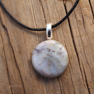 May include: A round, light-colored stone pendant with a silver bail hangs from a black cord. The stone has a mix of white, gray, and reddish-brown hues. The pendant is displayed on a rustic wooden surface.