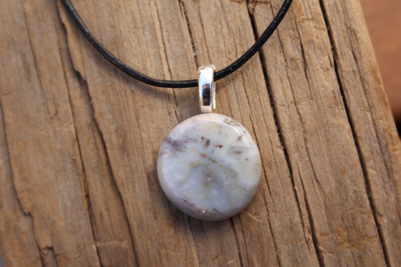 May include: A round, light-colored stone pendant with a silver bail hangs from a black cord. The stone has a mix of white, gray, and reddish-brown hues. The pendant is displayed on a rustic wooden surface.