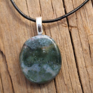 May include: A round pendant with a polished, mottled green and blue stone. The stone is attached to a silver-colored bail and hangs from a black cord. The pendant rests on a weathered wooden surface.