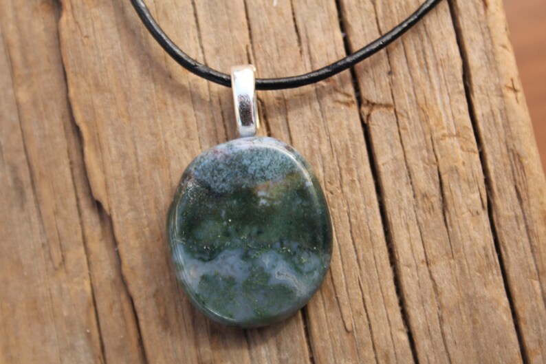 May include: A round pendant with a polished, mottled green and blue stone. The stone is attached to a silver-colored bail and hangs from a black cord. The pendant rests on a weathered wooden surface.