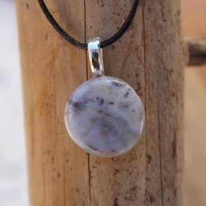 May include: A round pendant with a marbled, light blue and white stone. The pendant is suspended from a black cord and has a silver-colored metal bail. The background is a wooden surface.