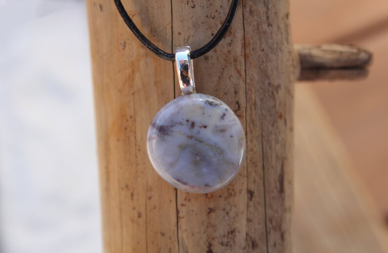 May include: A round pendant with a marbled, light blue and white stone. The pendant is suspended from a black cord and has a silver-colored metal bail. The background is a wooden surface.