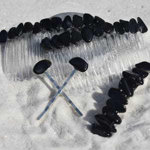 Könnte beinhalten: Eine Auswahl an Haaraccessoires, darunter ein durchsichtiger Plastikkamm mit schwarzen Steinen, zwei silberne Haarnadeln mit schwarzen Steinakzenten und eine Haarspange aus schwarzem Stein. Die Artikel sind auf einer weißen, sandigen Oberfläche angeordnet.