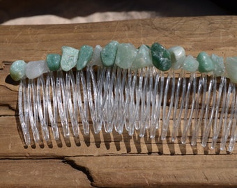 May include: A clear plastic hair comb adorned with a row of polished, irregularly shaped green and white gemstones. The comb's teeth are evenly spaced and create a contrast with the natural stone accents.
