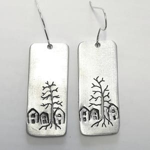 Puede incluir: Un par de pendientes de plata con un diseño rectangular que presenta un árbol y tres casas. Los pendientes tienen un cierre de gancho simple.