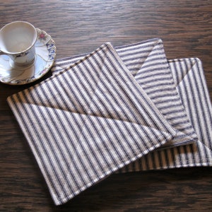 May include: Three square fabric coasters with a blue and white striped pattern are stacked on a wooden surface. A small white teacup with a gold handle and a floral patterned saucer sits to the left of the coasters.