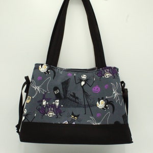 Op de afbeelding: Een grijze tote bag met zwarte hengsels en een zwarte bodem. De tas heeft een print van personages uit de film Nightmare Before Christmas, waaronder Jack Skellington, Sally en Zero. De print bevat ook pompoenen, vleermuizen en andere Halloween-themabeelden.