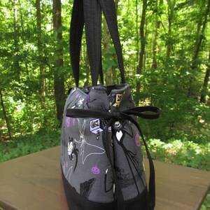 Op de afbeelding: Een zwarte tote bag met een grijze voering van stof met een herhalend patroon van personages uit The Nightmare Before Christmas. De tas heeft twee zwarte banden en een zwarte trekkoord sluiting.