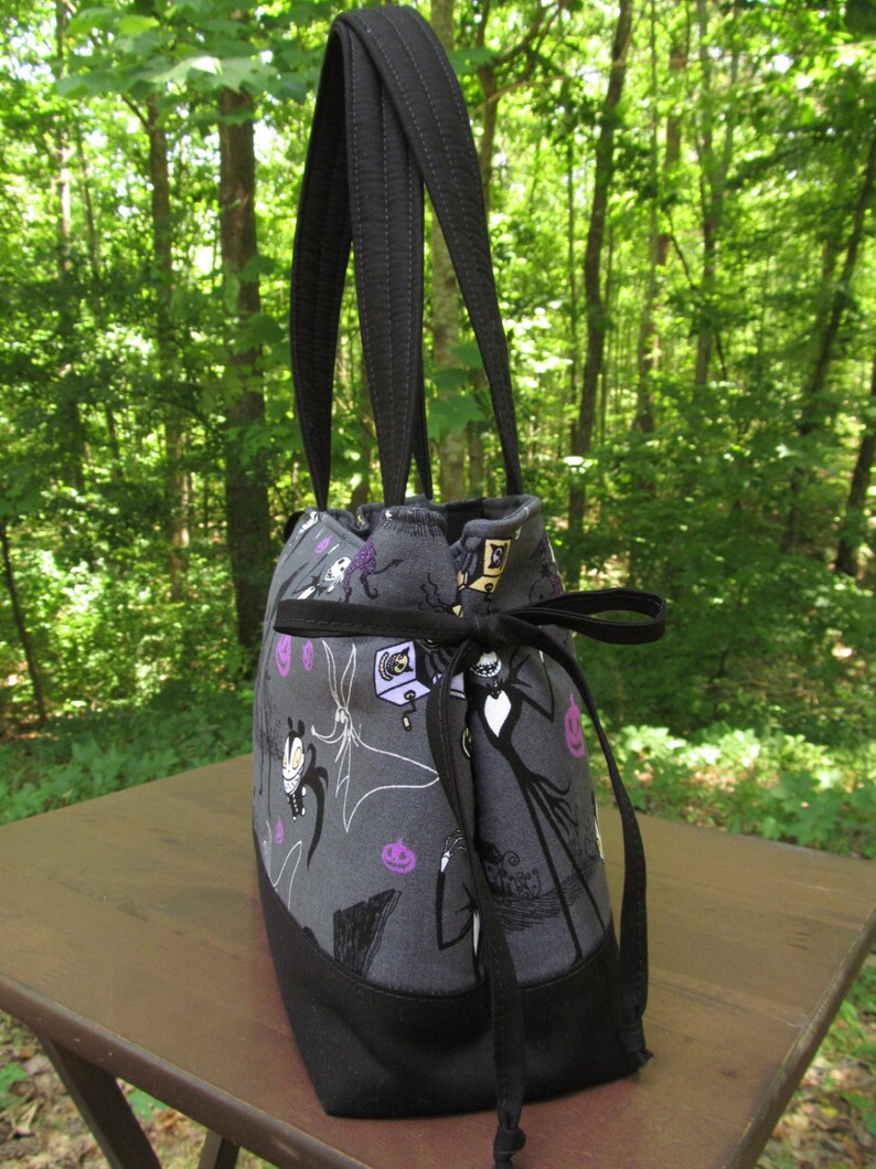 Op de afbeelding: Een zwarte tote bag met een grijze voering van stof met een herhalend patroon van personages uit The Nightmare Before Christmas. De tas heeft twee zwarte banden en een zwarte trekkoord sluiting.