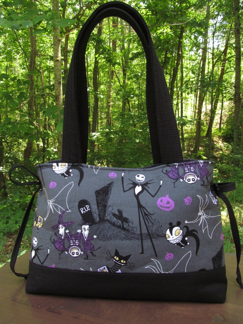 Op de afbeelding: Een zwarte tote bag met een grijze stof met een zwart-witte illustratie van personages uit The Nightmare Before Christmas. De personages zijn Jack Skellington, Sally en Oogie Boogie. De tas heeft een zwarte band en een zwarte bodem.